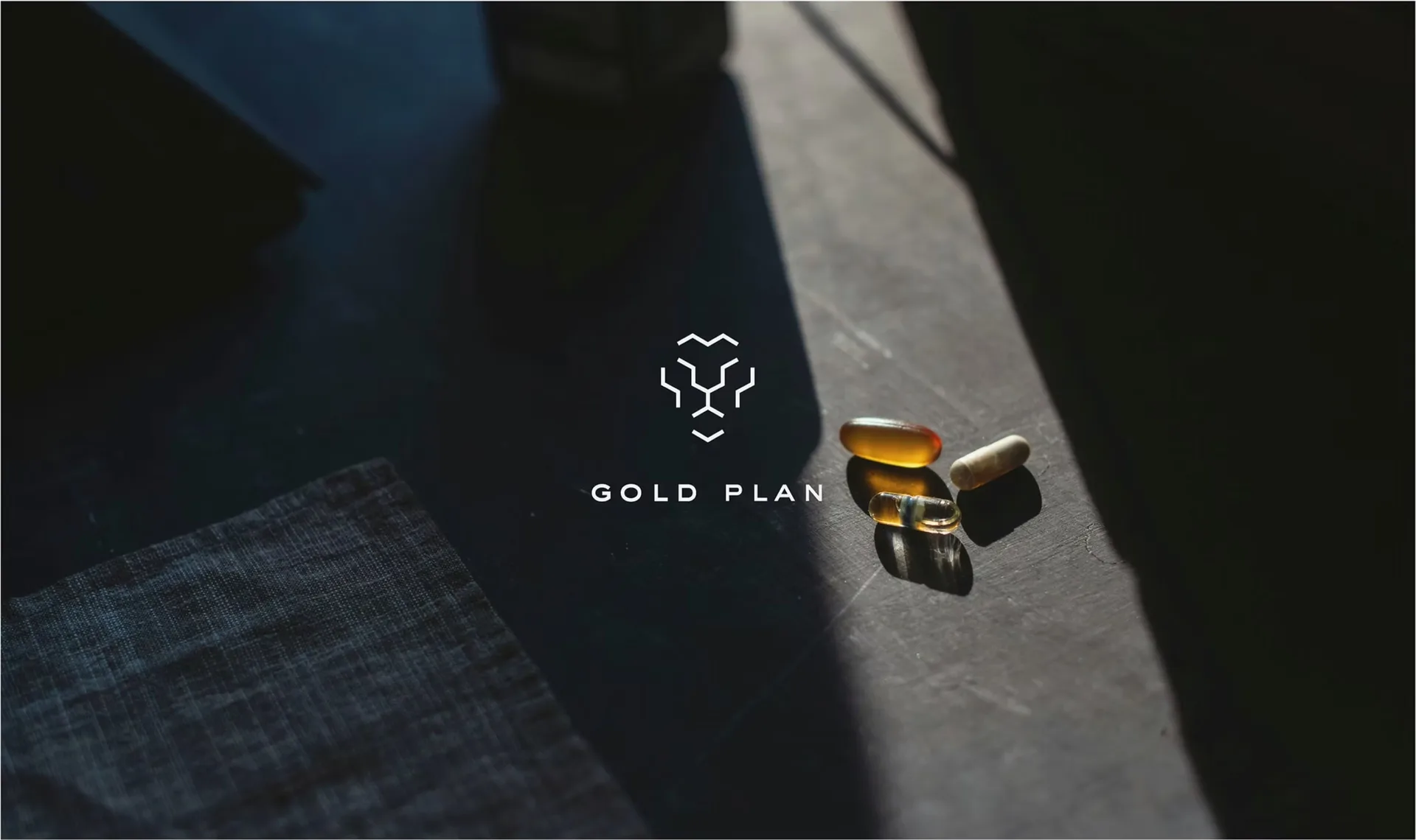 Logotipo Gold Plan junto a cápsulas doradas sobre una mesa en penumbra